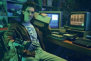 A young white man in glasses and slicked back 80s mullet wearing a "power glove" and wearing t t-shirt and worn brown leather jacket with jeans holds a large oldfashioned keyboard on one shoulder and sits in front of two old computers and technical clutter