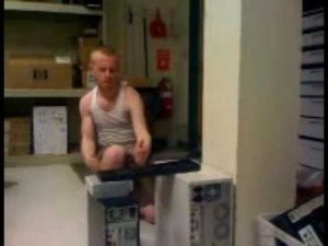 Young white man in cluttered garage about to karate chop a keyboard laying across two computer boxes.